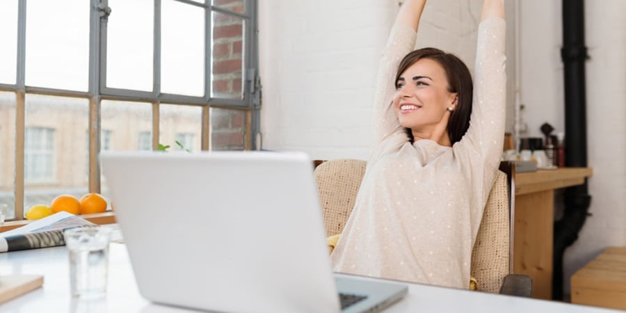 Happy relaxed young woman sitting in her kitchen with a laptop in front of her stretching her arms above her head and looking out of the window with a smile