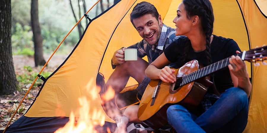 Your couple sitting outside tent by bonfire playing guitar and drinking coffee